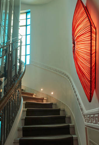 Elegant staircase with marble steps, glass elevator, soft wall lighting, and a striking red diamond-shaped wall light, accented by a tall window with colored glass panels