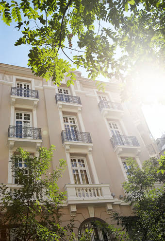 Sun-dappled façade of The Excelsior Hotel, with ornate balconies and tall windows, framed by lush greenery and bright natural light for a fresh, welcoming atmosphere