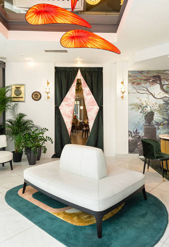 Bright hotel lobby with modern white seating, vibrant orange sculptural ceiling lights, lush green plants, a unique curved reception desk, and a large botanical mural, creating a welcoming and stylish atmosphere with views to the outdoor café terrace