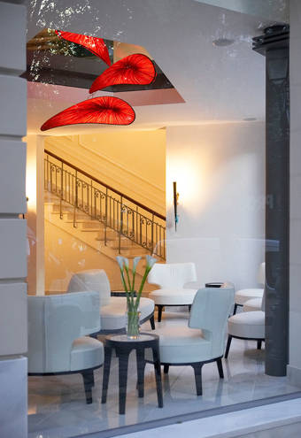 Lobby view through a large window, showcasing modern white armchairs, a small table with calla lilies, striking red sculptural ceiling lights, and an elegant staircase with a decorative railing in a bright, welcoming setting