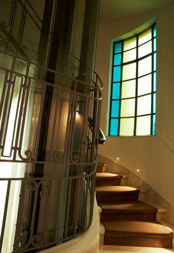 Elegant public indoor view featuring a curved staircase with soft carpeting, an art deco glass elevator, and a large window with colorful blue and yellow panels, creating a refined, welcoming atmosphere